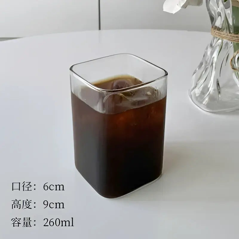 A clear minimalist square glass juice cup filled with dark iced coffee for kitchen drinkware use
