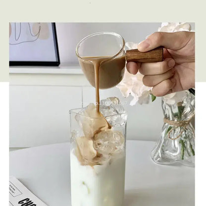 A clear glass juice cup with wooden handle filled with coffee being poured into an iced drink square glass juice cup