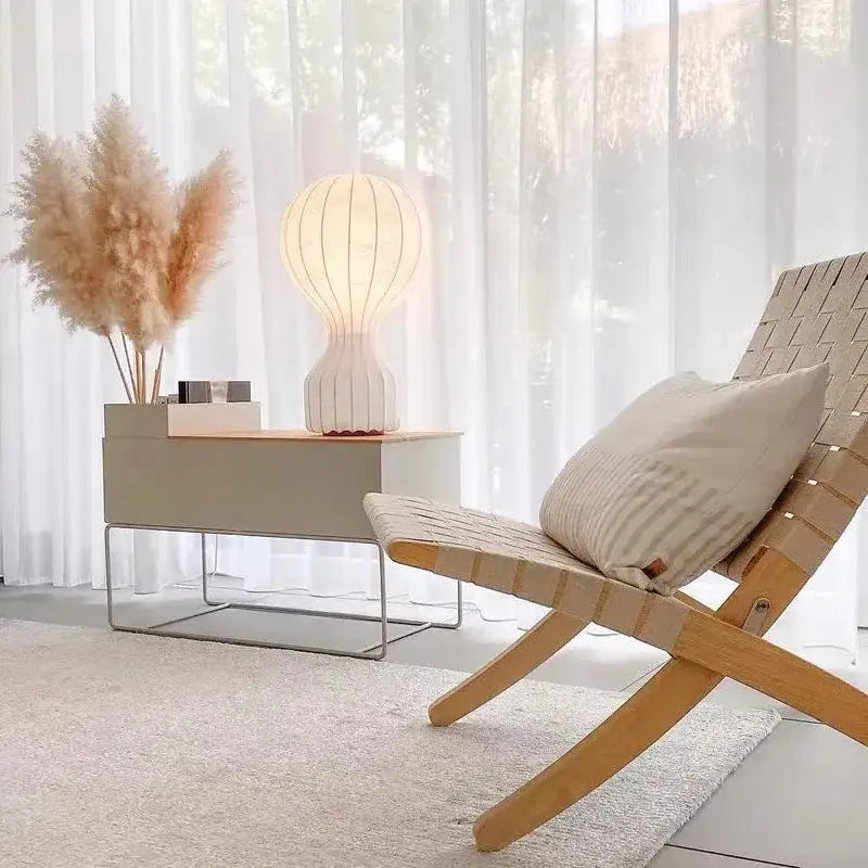 Light beige woven wood lounge chair with striped cushion shown beside minimalist white ribbed gatto cocoon silk table lamp