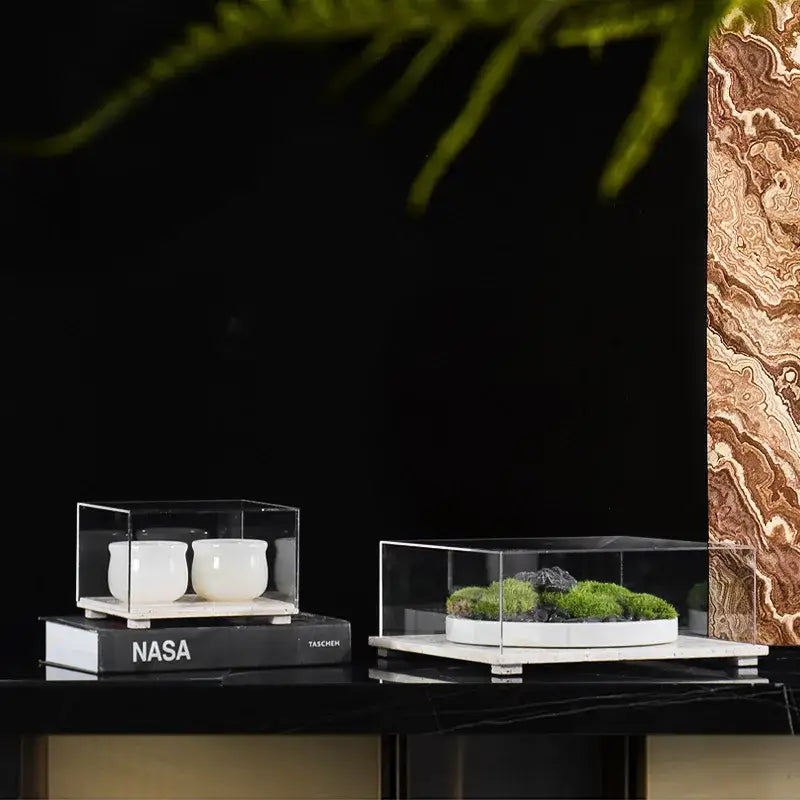 Two clear acrylic display cases with travertine base, one holding white ceramic cups and the other green moss with stones