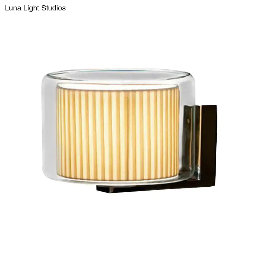 Modern wall lamp with a cylindrical clear glass casing and warm amber pleated fabric shade for bedroom lighting