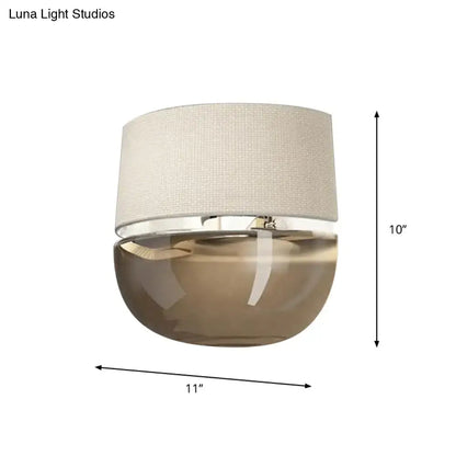 Modern wall sconce with textured beige fabric shade and tan glass amber base with polished chrome trim for bedroom lighting