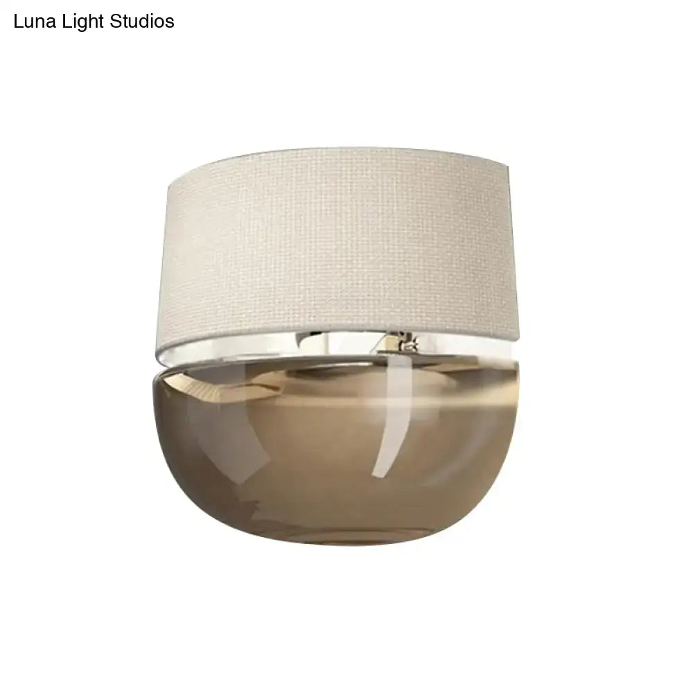 Modern wall sconce with textured beige fabric shade and tan glass smoked base accented by polished chrome trim for bedroom