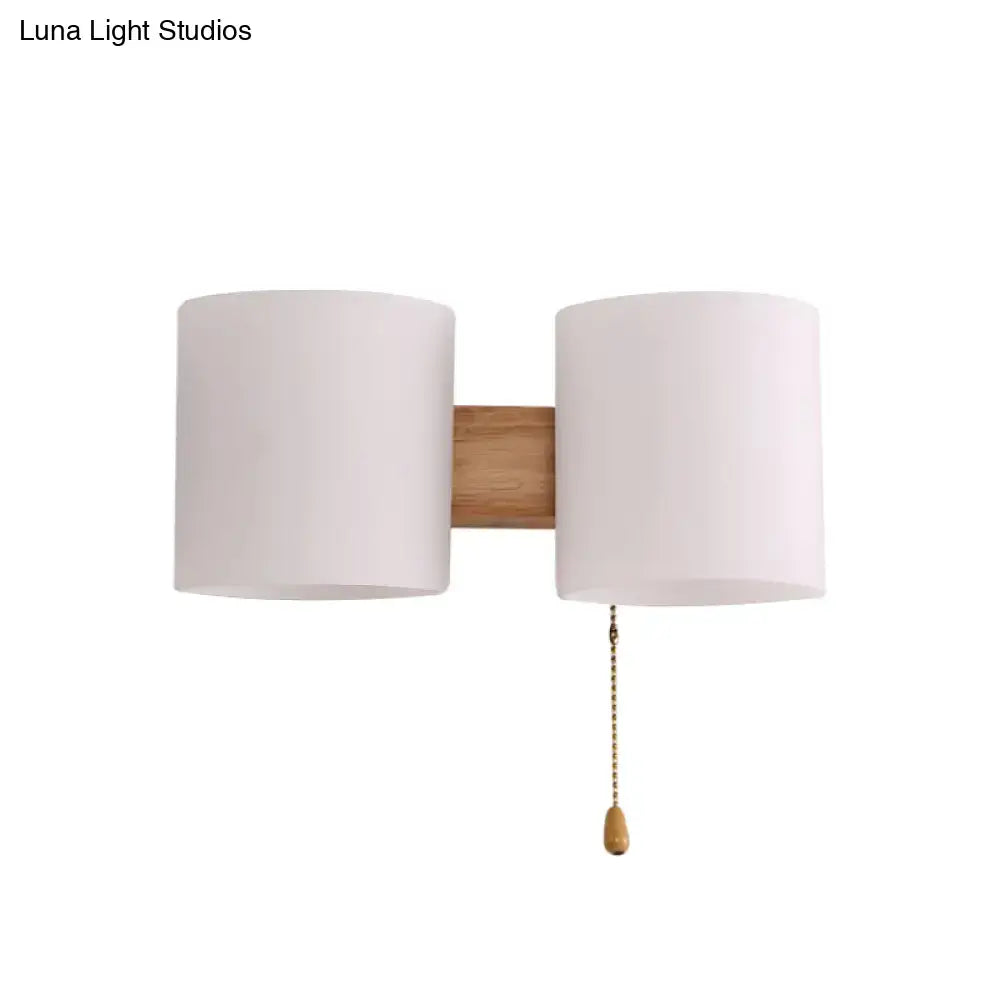 Modern beige fabric and wood wall light bedroom sconce with two cylindrical shades and brass pull chain on natural wood bar