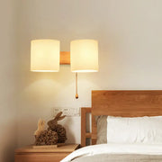 Modern beige fabric and wood wall light bedroom sconce with warm-toned cylindrical lampshade and pull chain switch