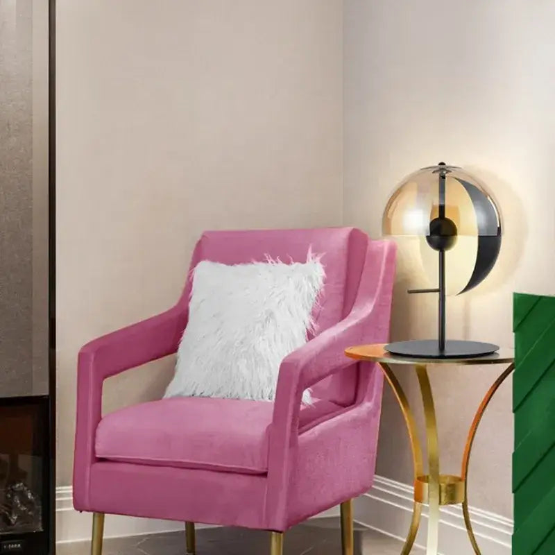 A plush pink velvet armchair with gold tone legs and white faux fur pillow beside a modern black glass designer bedside lamp