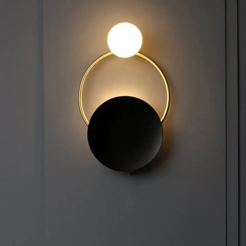Modern black and gold metal planet wall mount lamp with glowing white spherical bulb and matte black disc bedroom sconce
