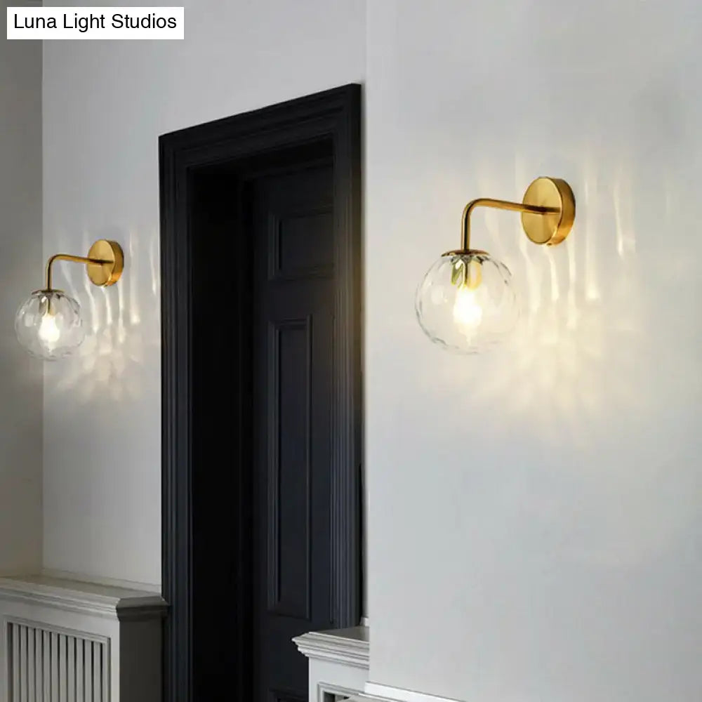 Gold-finished wall mount light with clear textured glass globe and modern black metal rippled glass bedroom sconce design