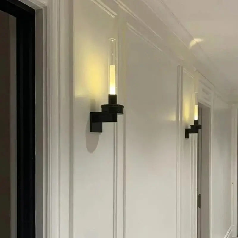 Modern black metal wall light with clear glass cylinder housing an illuminated led candle-style bulb for bedroom use