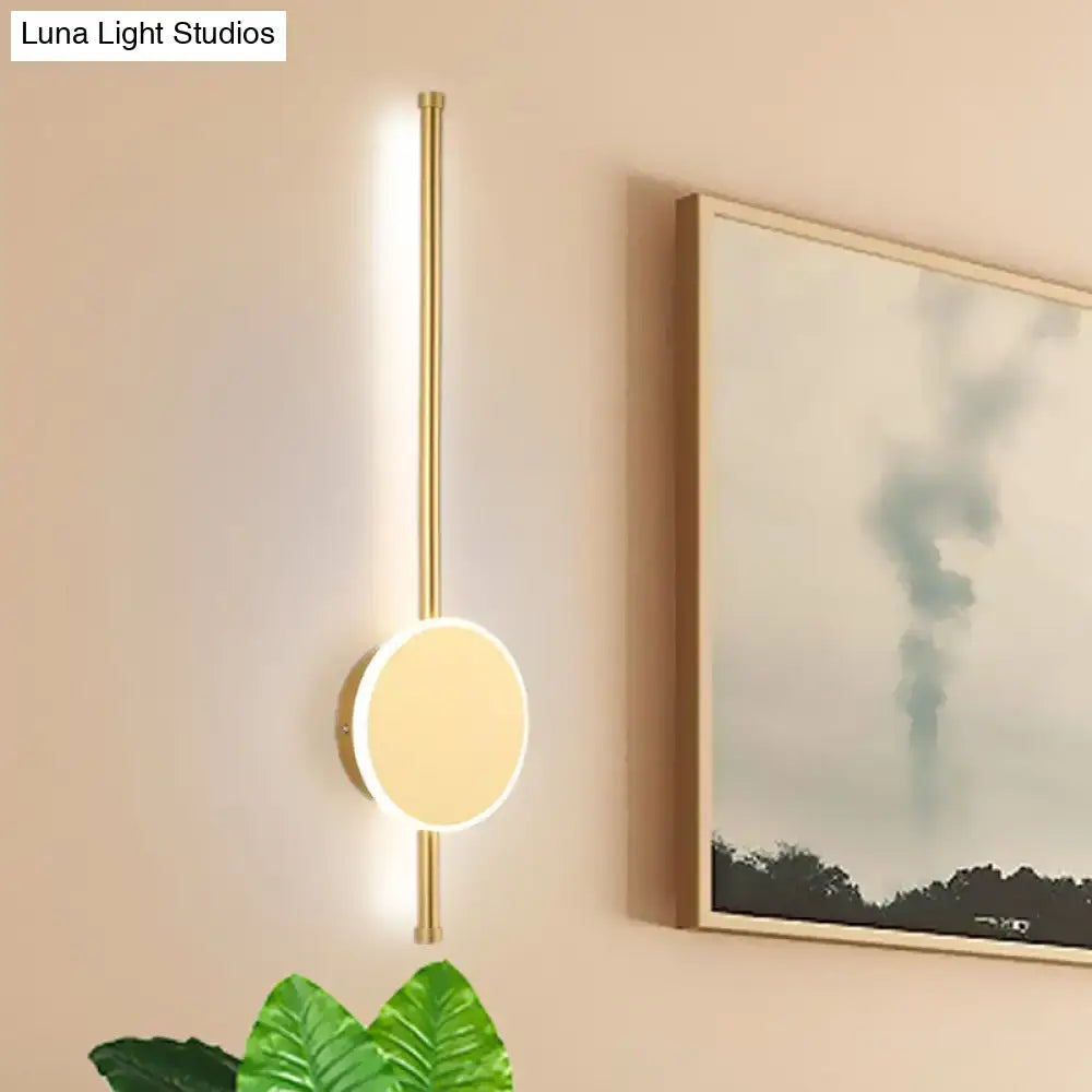 Gold-toned minimalist wall sconce with sleek vertical rod and circular illuminated face featuring warm color temperature