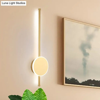 Gold-toned minimalist wall sconce with sleek vertical rod and circular illuminated face featuring warm color temperature