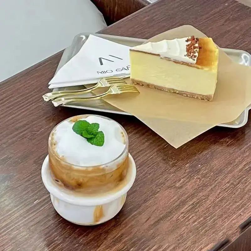 A creamy yellow cheesecake slice on a parchment tray beside a modern borosilicate cap glass coffee tumbler cup