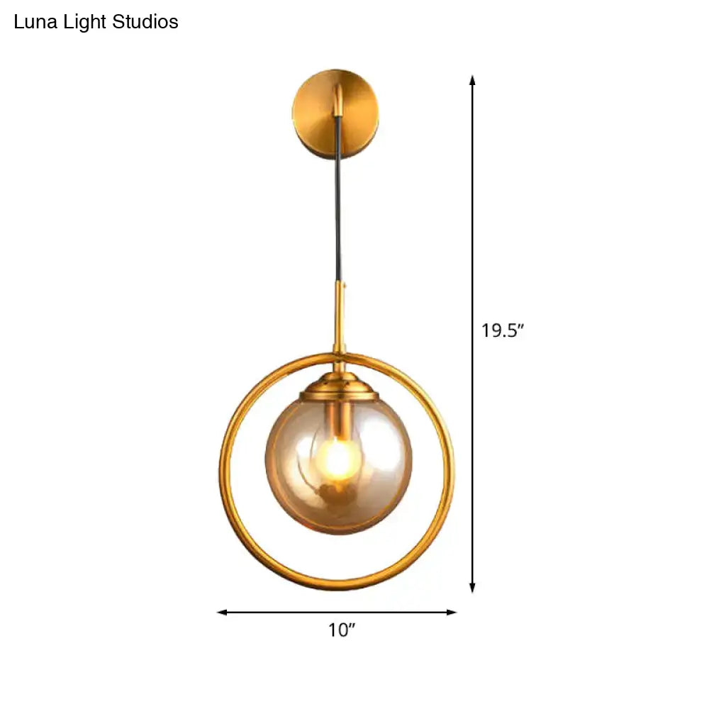 Modern brass circular wall mounted lamp with a gold-toned frame and amber glass globe shade for bedroom use