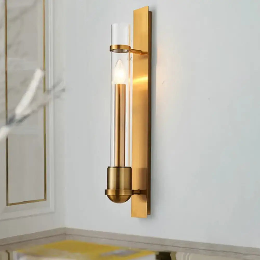 Modern brass clear glass wall sconce light fixture with gold-toned frame and illuminated candle-style bulb for bedroom use