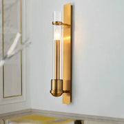 Modern brass clear glass wall sconce light fixture with gold-toned frame and illuminated candle-style bulb for bedroom use