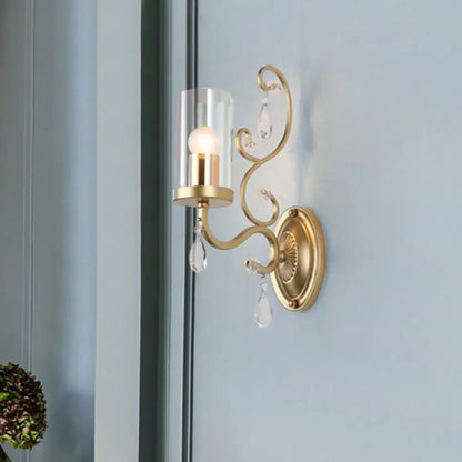 Modern brass crystal wall mounted light with gold-toned sconce, clear crystal droplets, and cylindrical glass shade