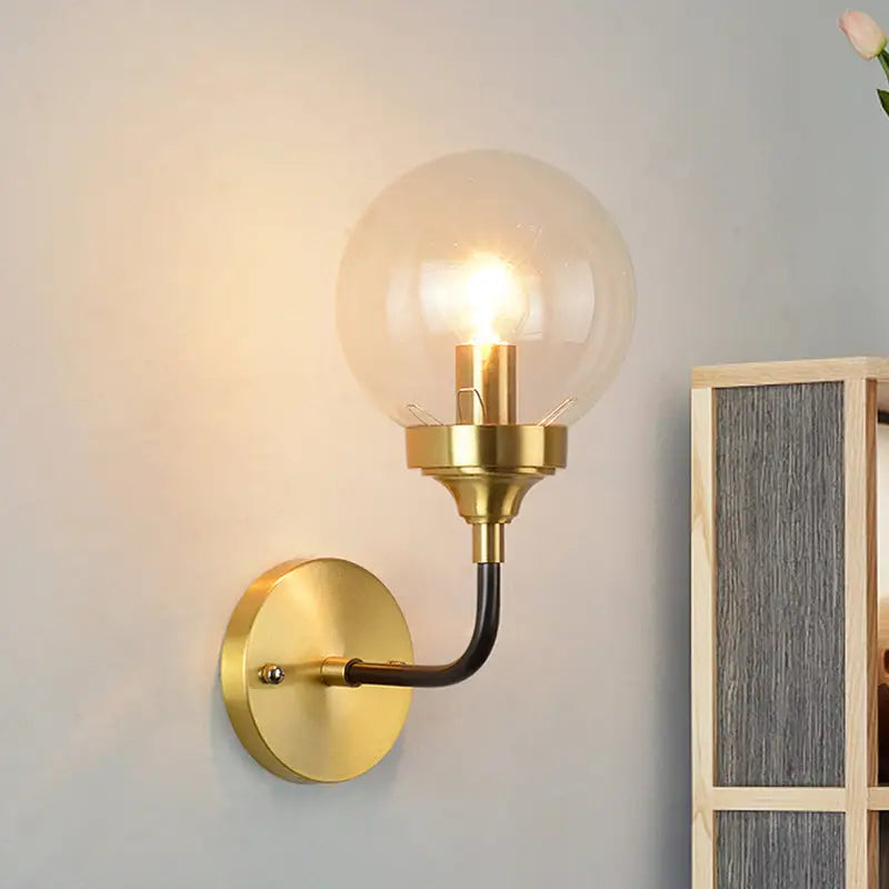Modern brass curved arm wall sconce with clear glass globe and illuminated bulb for bedroom lighting