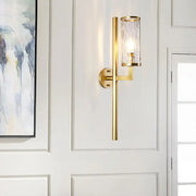 Modern brass cylinder sconce with clear water glass textured cylinder and sleek gold-toned metal arm bedroom lamp