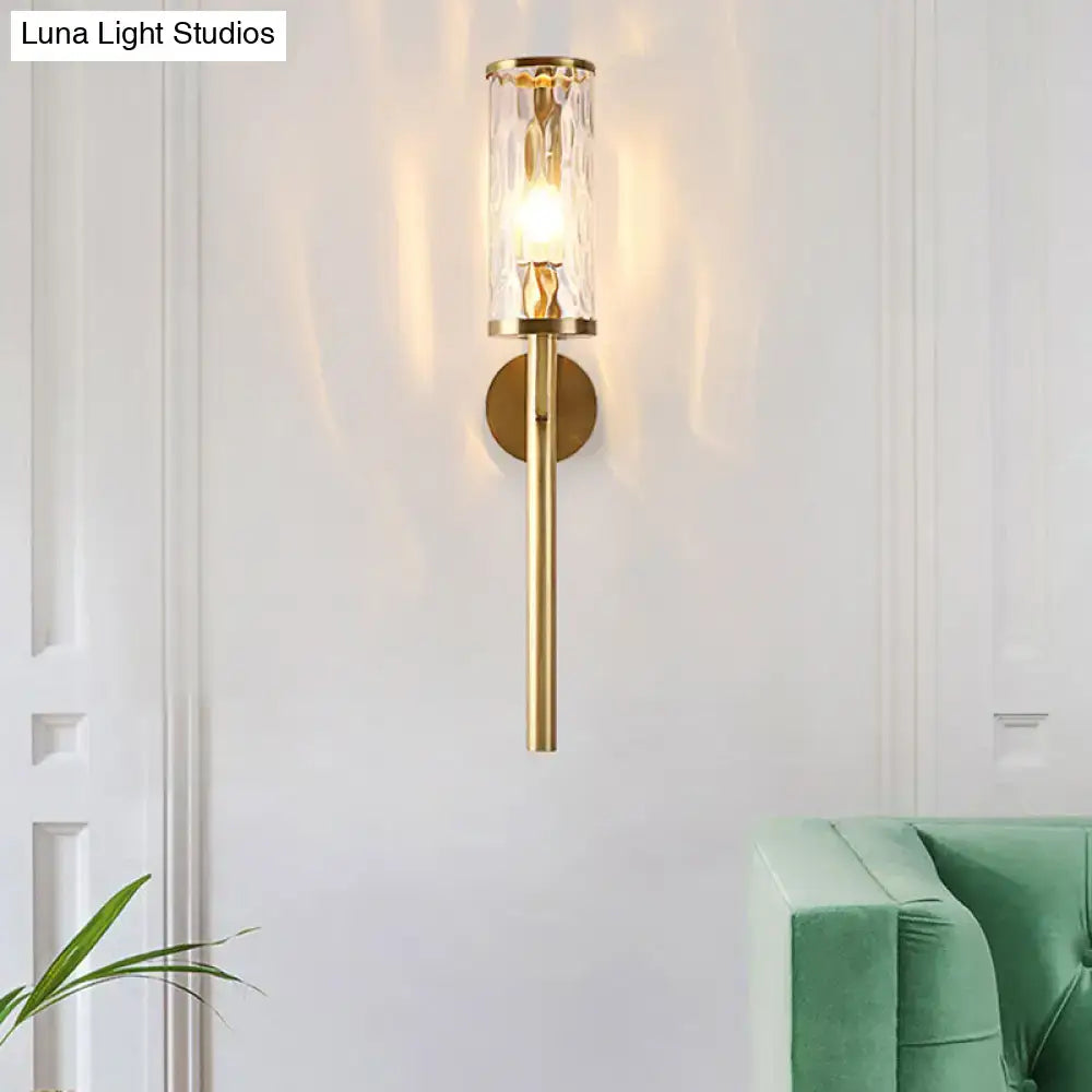 A modern brass cylinder sconce with clear water glass textured cylinder housing an illuminated bulb for bedroom use