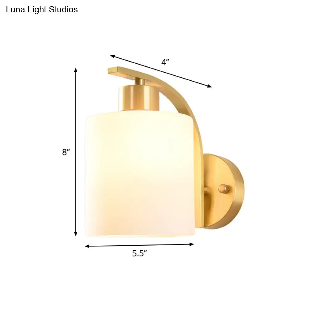 Modern brass cylinder wall sconce with gold finish and milk glass shade featuring an arched arm for bedroom lighting