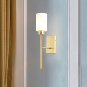 Modern brass wall light with a gold-toned frame and cylindrical opal glass tube shade for bedroom use
