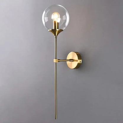 Modern brass pencil arm wall light with gold-toned finish, clear glass globe, and exposed bulb for bedroom use