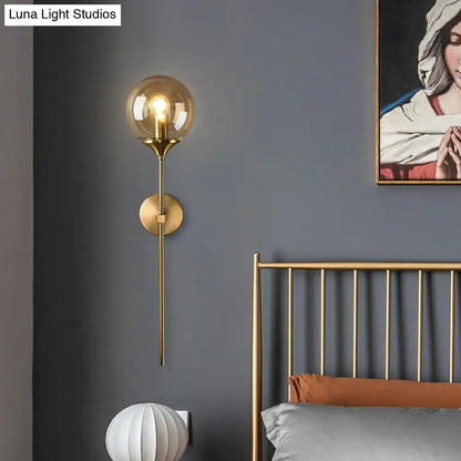 Modern brass pencil arm wall light with gold-toned spherical glass shade and slender metallic stem for bedroom use
