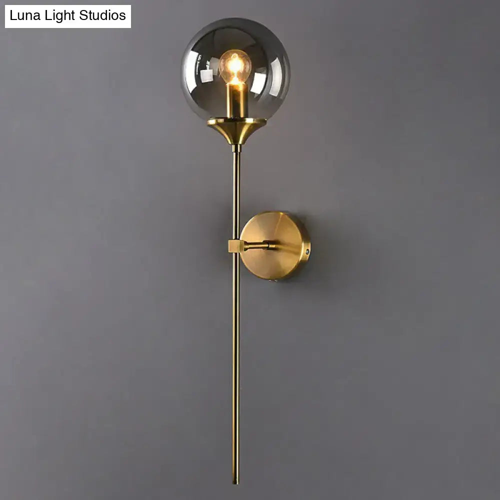 A modern brass pencil arm wall light featuring a smoked glass globe and exposed filament bulb for bedroom use