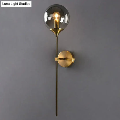 A modern brass pencil arm wall light featuring a smoked glass globe and exposed filament bulb for bedroom use
