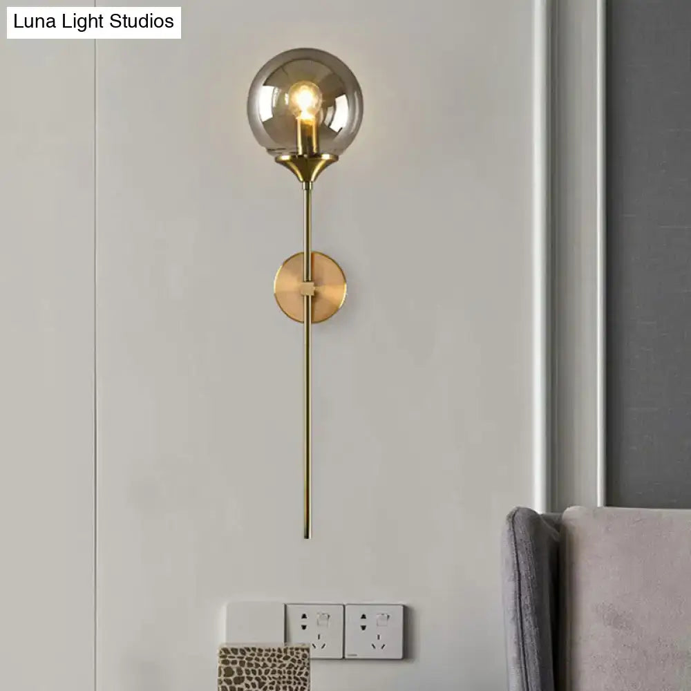 Modern brass pencil arm wall light featuring a smoke gray glass globe and brass metal stem for bedroom use