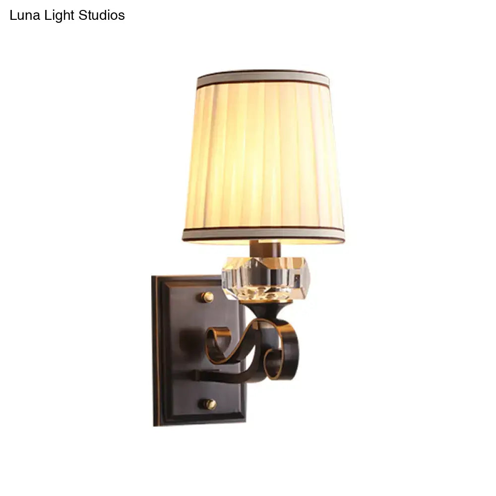 A warm-toned pleated shade on a black brass direct wired electric wall lamp with a dark bronze ornate arm and crystal accent