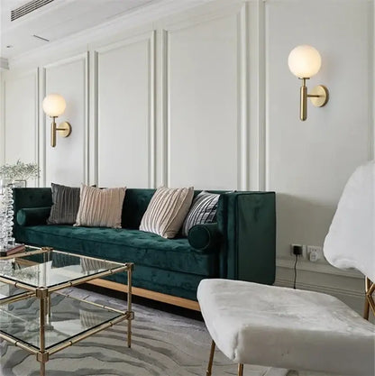 Modern brass wall lights with white and clear decorbites glass ball mounted above a deep emerald green velvet sofa with tufted backrest