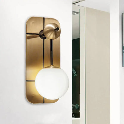 A modern brass wall sconce with a brushed brass backplate and opal glass bulb for direct wired bedroom lighting