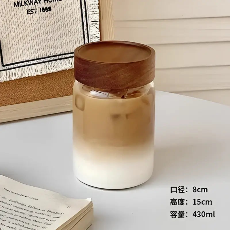 Modern clear glass tea cup with wooden lid filled with iced coffee showing brown and white gradient layers