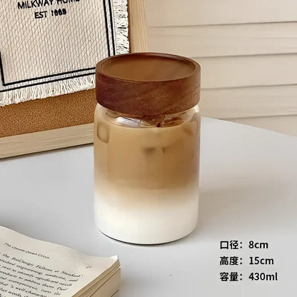 Modern clear glass tea cup with wooden lid filled with iced coffee showing brown and white gradient layers