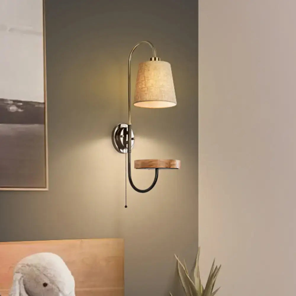 Modern conical fabric wall mount lamp with beige shade, pull chain, curved metallic arm, and wooden accent shelf for bedroom lighting