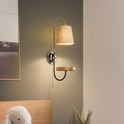 Modern conical fabric wall mount lamp with beige shade, pull chain, curved metallic arm, and wooden accent shelf for bedroom lighting