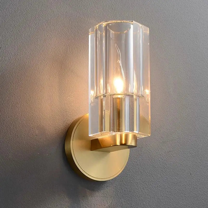 A modern decorbites crystal wall lamp featuring a clear faceted glass cylinder and gold metal base for bedroom decor