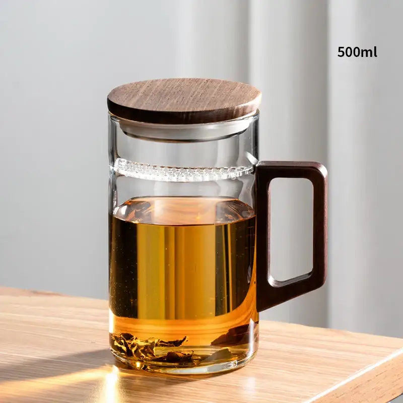 Clear glass teapot with dark wood lid and handle filled with amber tea in modern dark green glass tea cup set