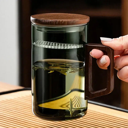 A clear glass teapot with dark wood lid and handle showcasing steeping tea leaves in a modern dark green tea cup set