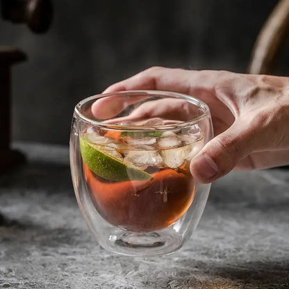 Modern double walled transparent glass cup holding amber liquid with ice and lime wedge in kitchen and dining room use