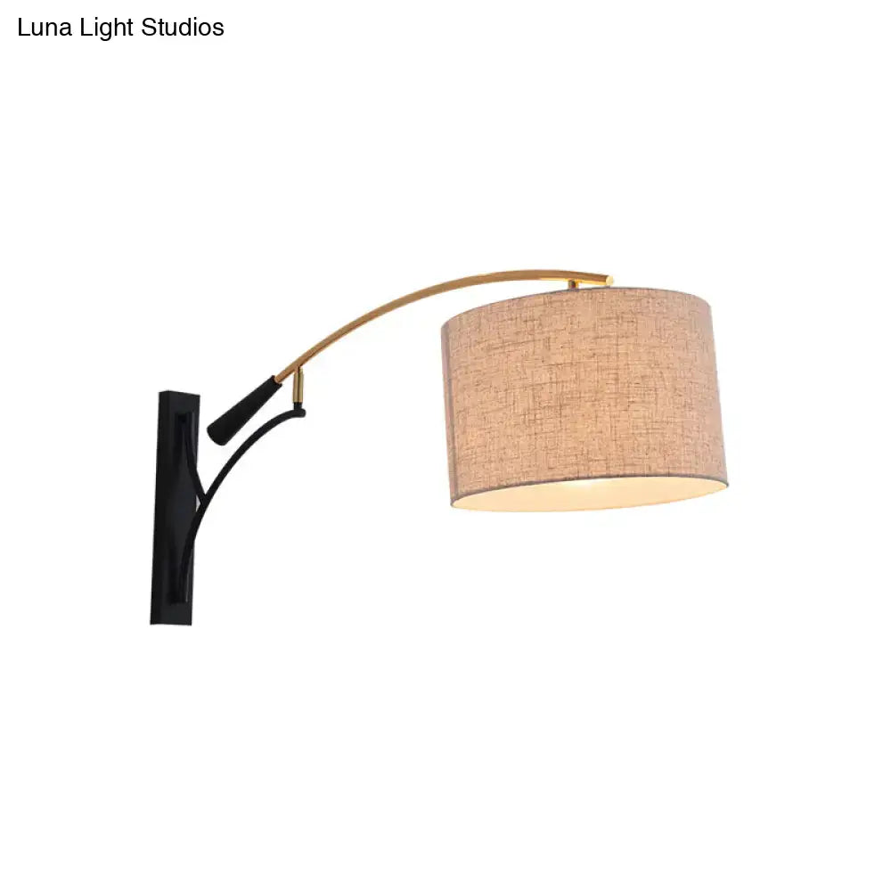 A modern drum shade wall lamp with a curved brass arm, black bracket, and flaxen coffee fabric shade for bedroom