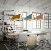 Three modern geometric book pendant lamps in mustard yellow teal blue and dusty rose pink hang above a wooden table