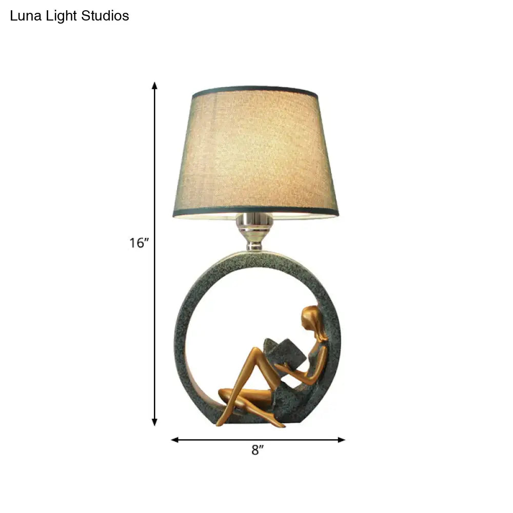 A bronze-colored modern girl reading resin night lamp with fabric shade and off-white circular base for bedroom decor