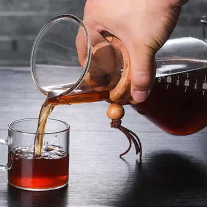 A clear glass coffee kettle with wooden handle and steel filter drip pouring dark amber liquid into matching glass cup