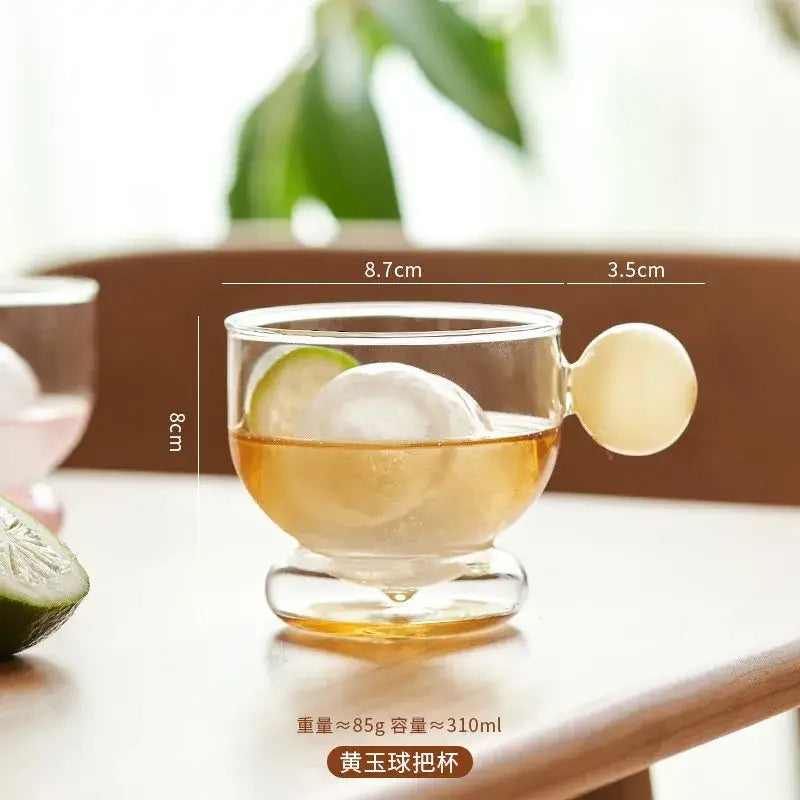 Clear glass coffee mug with a rounded translucent yellow glass spherical handle and a footed base for kitchen use