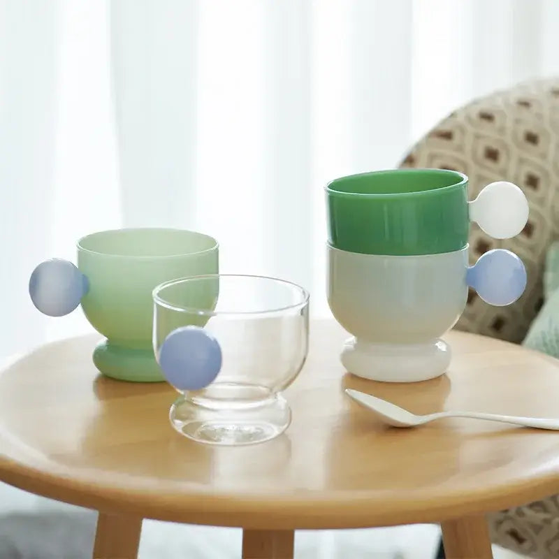 Pastel green and white ceramic coffee mugs with rounded blue spherical handle sit on wooden table for kitchen use