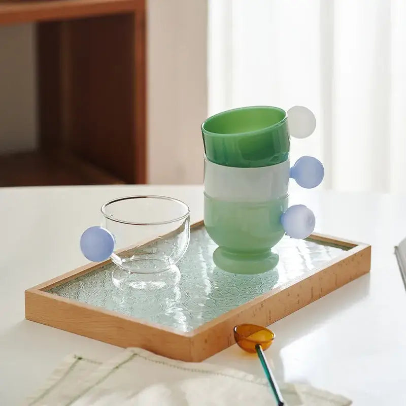Stacked modern green gradient glass coffee mugs with translucent blue spherical handles on a wooden tray