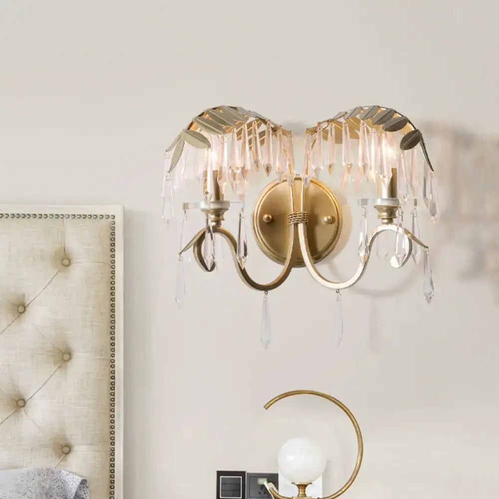 Modern gold wall sconce light with bent arm featuring leaf-shaped metal accents and crystal droplets for bedroom use