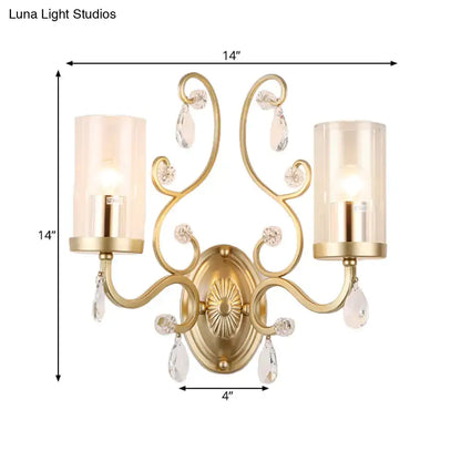 Gold-toned ornate wall light with two candle-style clear glass cylinder shades and dangling crystal teardrops for bedroom use